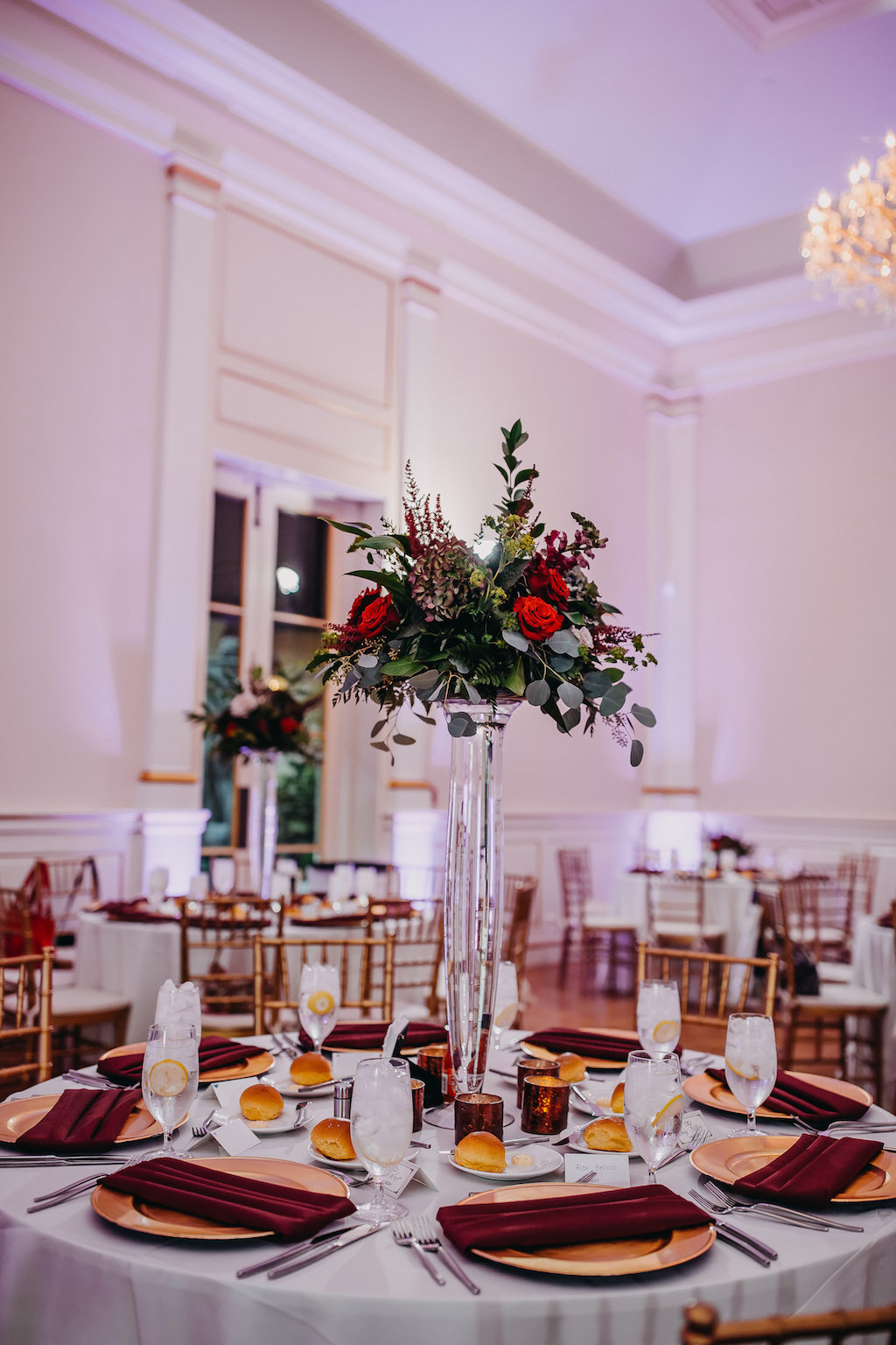 Modern Ballroom Wedding Reception Round Table with Red Linens, Red and Orange Floral with Greenery Tall Centerpiece in Clear Glass Vase, with Gold Chargers