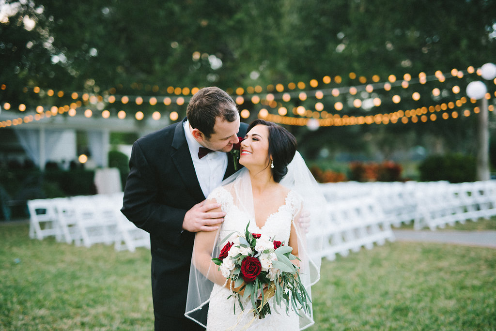 Outdoor Garden Wedding Ceremony Portrait, Bride with Red Rose, White Floral, and Greenery Bouquet, Groom with Burgundy Tie, with String Lights and White Folding Chairs | Tampa Wedding Photographer Kera Photography