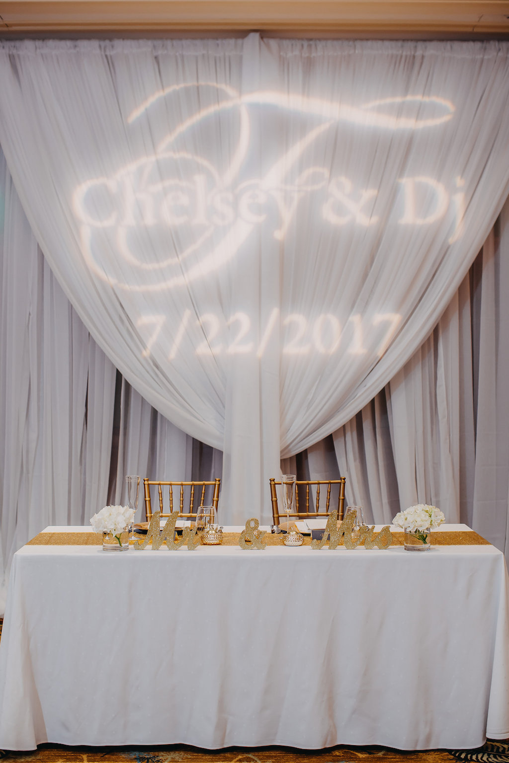 Custom Bride and Groom Name Light Projection on White Draping Behind Simple Sweetheart Table with Gold Mr and MRs Sign and Gold Table Runner | Tampa Bay Wedding Rentals and Lighting Gabro Event Services