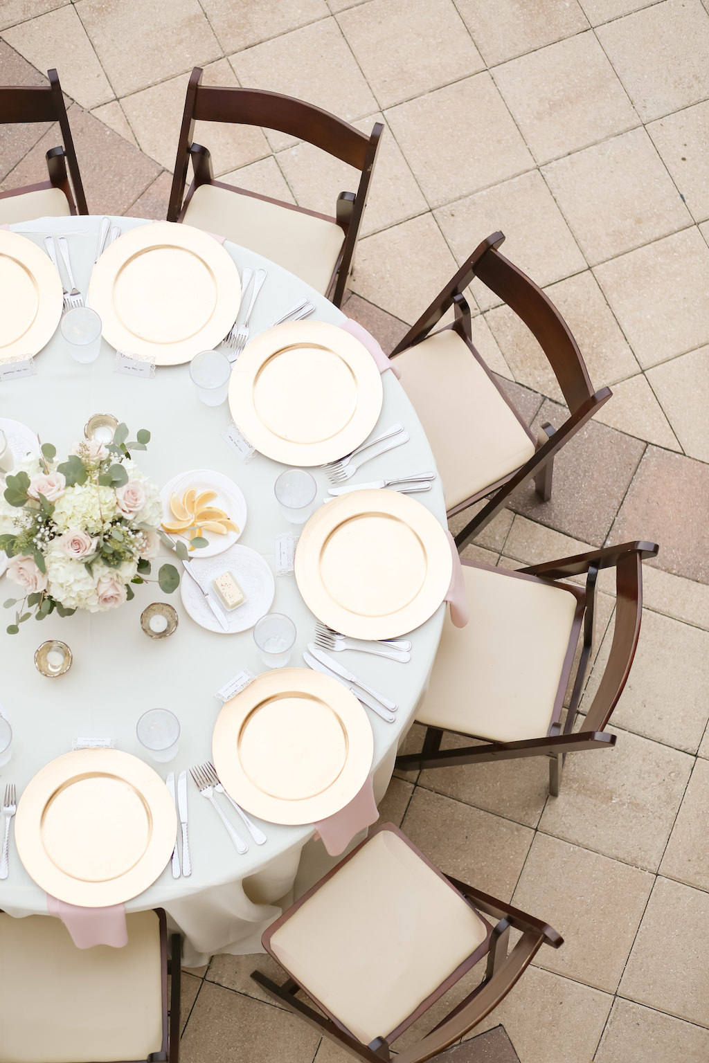 Outdoor Rustic Nautical Wedding Reception ROund Table Decore with Ivory Rose and Greenery Centerpiece, Blue Tablecloth and Blush Pink LInens, and Gold Chargers and Brown Wooden Chairs