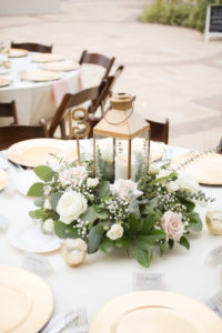 Rustic Nautical Outdoor Wedding Reception Round Table Decor with Gold Hurricane Lantern and Low White Rose with Greenery Centerpiece, Gold Chargers and Tea Candles, Light Blue Tablecloth and Pink Linens, and Brown Wood Folding Chairs