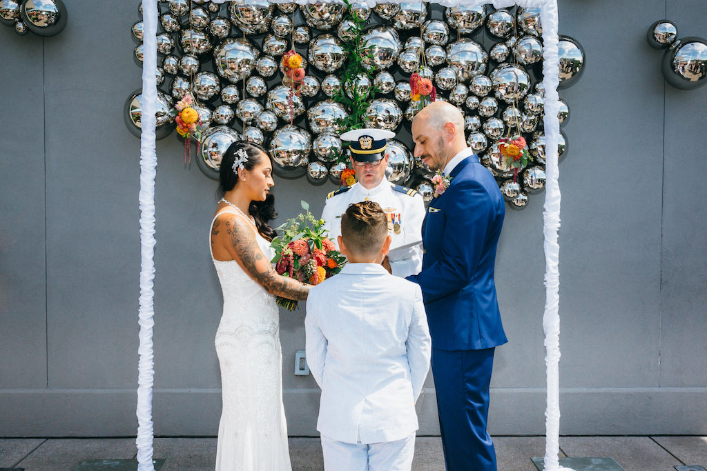 Outdoor Rooftop Wedding Ceremony Portrait with Simple Rectangular Arch, Silver Ball Wall Art, and Hanging Orange and Green Florals and Bouquet, Groom in Blue Suit | Tampa Hotel Wedding Venue The Epicurean | Planner UNIQUE Weddings and Events