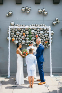 Outdoor Rooftop Wedding Ceremony Portrait with Simple Rectangular Arch, Silver Ball Wall Art, and Hanging Orange and Green Florals and Bouquet, Groom in Blue Suit | Tampa Hotel Wedding Venue The Epicurean | Planner UNIQUE Weddings and Events