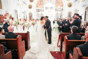 Greek Wedding Ceremony Kiss Portrait with Bridesmaids in Floorlength White Dresses, Groomsmen in Black Tuxes with White Boutonniere | Tarpon Springs Traditional Wedding Venue St. Nicholas Greek Orthodox