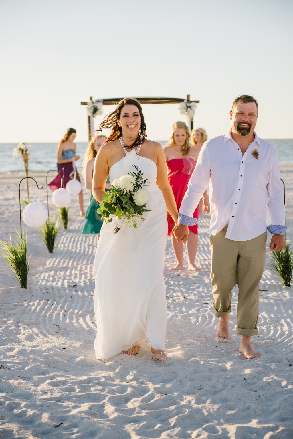Simple Intimate Beach Wedding Ceremony Portrait with White Pomander Balls on Iron Hook with Greenery, Wooden Arch with White Flowers and Draping, and Tiki Torches, Bridesmaids in Mismatched Bright Colored Tropical Dresses, Bride in Halter DaVinci Dress | Tampa Bay Wedding Planner Gulf Beach Weddings | Treasure Island Venue Sunset Vista Condo Hotel Resort 