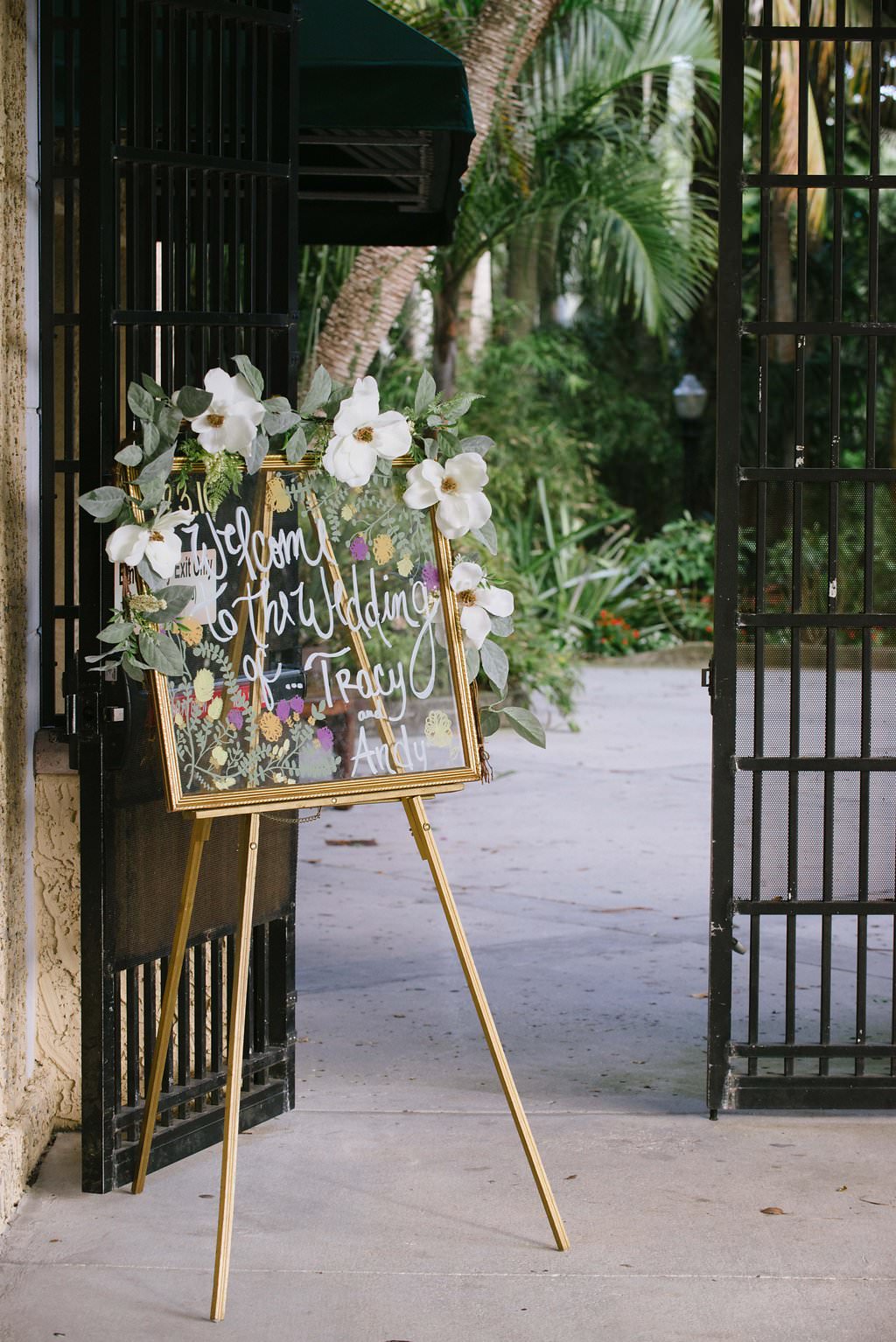 Outdoor Garden Wedding Welcome Sign, Handwritten on Glass in Gold Frame with Large White Flowers and Greenery