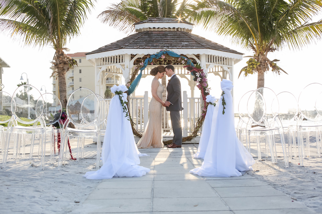 Outdoor Beach Wedding Ceremony at Gazebo with Clear Plastic Chairs, White Draping, Whimsical Purple, Black, Magenta, Fuchsia, Plum and White Floral Wedding Arch with Greenery with Turquoise Draping | Tampa Bay Wedding Florist Gabro Event Services | Waterfront Venue Isla Del Sol Yacht and Country Club | Planner Kelly Kennedy Weddings and Events | Photographer Lifelong Studios Photography