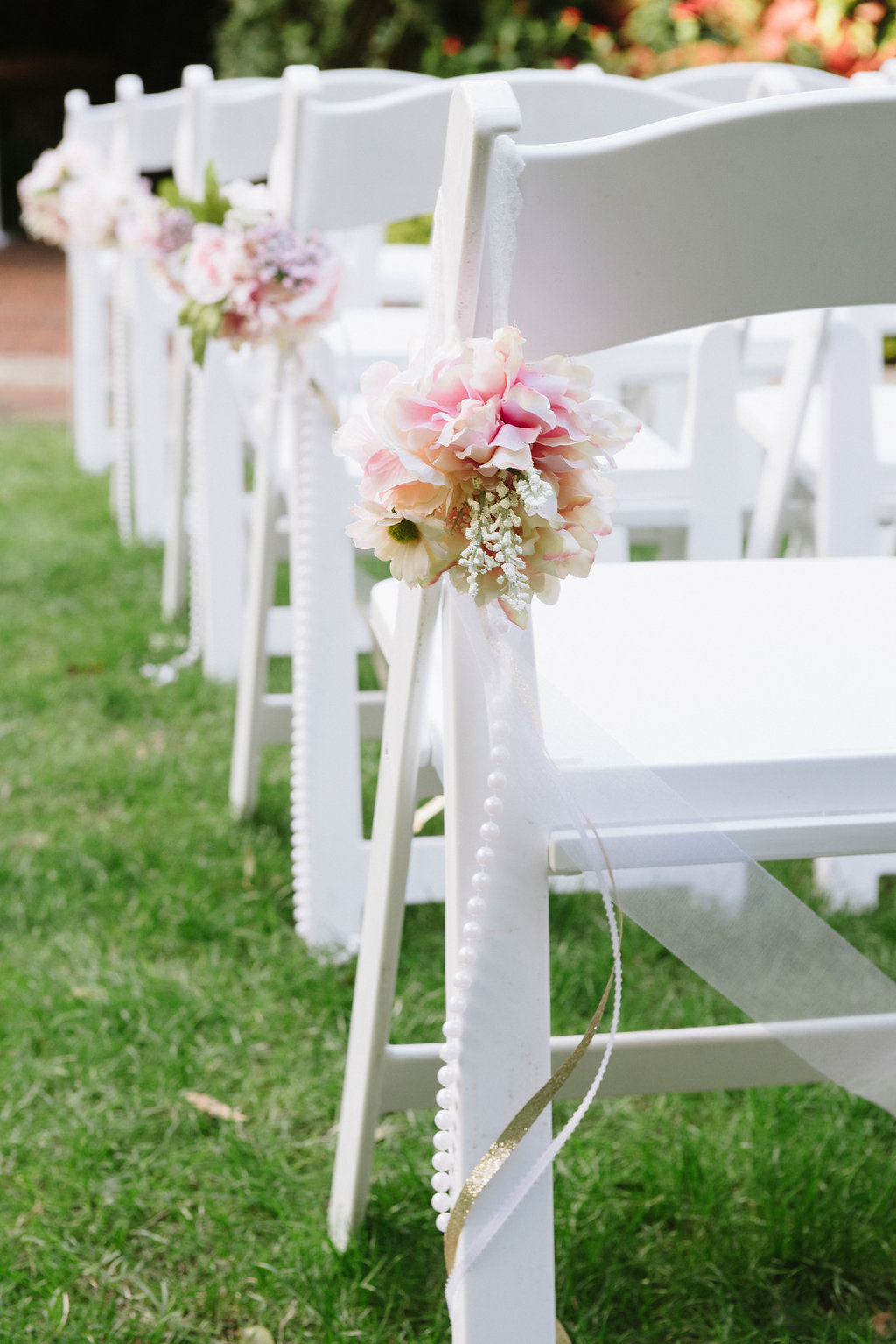 Outdoor Garden Wedding Ceremony Decor with White Folding Chairs with Pink and White Flowers, Pearl Beads, and Gold Ribbon