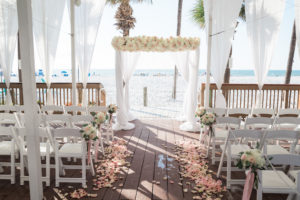 Outdoor Beachfront Sandpiper Deck Wedding Ceremony with White Folding Chairs, Blush Pink Rose Petal Aisle, White Floral with Greenery and Pink Ribbon, and White Draped Chuppah Wedding Arch with White and Pink Floral | Tampa Bay Event Draping and Rentals Gabro Event Services | Clearwater Beach Wedding Venue Hilton Clearwater Beach