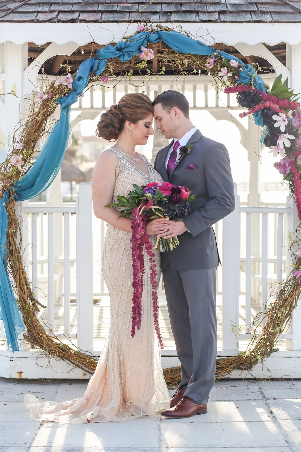 Outdoor Beach Wedding Ceremony at Gazebo with Whimsical Purple, Black, Magenta, Fuchsia, Plum and White Floral Wedding Arch with Greenery with Turquoise Draping, Bride in Jeweled Ivory Illusion Neckline Dress with Cascading Bouquet, Groom in Gray Suit with Fuchsia Tie | Tampa Bay Wedding Florist Gabro Event Services | Waterfront Venue Isla Del Sol Yacht and Country Club | Dress Shop Truly Forever Bridal | Menswear Sacino's Formalwear | Photographer Lifelong Studios Photography