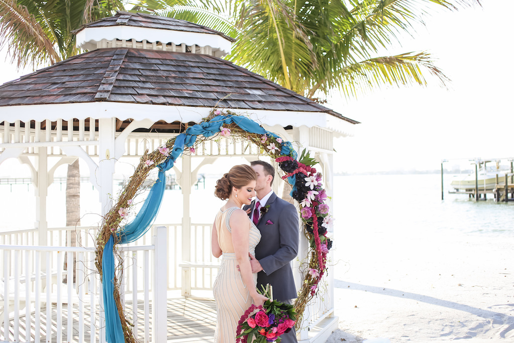 Outdoor Beach Wedding Ceremony at Gazebo with Whimsical Purple, Black, Magenta, Fuchsia, Plum and White Floral Wedding Arch with Greenery with Turquoise Draping, Bride in Jeweled Ivory Illusion Neckline Dress with Cascading Bouquet, Groom in Gray Suit with Fuchsia Tie | Tampa Bay Wedding Florist Gabro Event Services | Waterfront Venue Isla Del Sol Yacht and Country Club | Dress Shop Truly Forever Bridal | Menswear Sacino's Formalwear | Photographer Lifelong Studios Photography