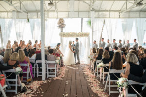 Outdoor Beachfront Sandpiper Deck Wedding Ceremony Portrait with White Folding Chairs, Blush Pink Rose Petal Aisle, White Floral with Greenery and Pink Ribbon, and White Draped Chuppah Wedding Arch with White and Pink Floral, Groomsmen in Light Gray Suits, Bridesmaids in Pink | Tampa Bay Event Draping and Rentals Gabro Event Services | Clearwater Beach Wedding Venue Hilton Clearwater Beach