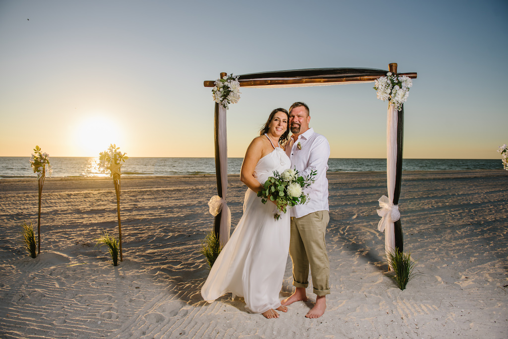 Outdoor Sunset Beach Wedding Ceremony Portrait with White Draped Wooden Ceremony Arch with Flowers and Greenery, Bride in Halter DaVinci Dress with White Floral Bouquet with Natural Greenery | Tampa Bay Wedding Planner Gulf Beach Weddings | Treasure Island Wedding Venue Sunset Vista Condo Hotel Resort 