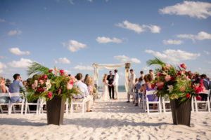 Outdoor Beach Wedding Ceremony Portrait with Bamboo Ceremony Arch With Wrapped White Draping, and Tall Blush, Pink, Magenta Rose, White Lilly, and Palm Frond Tropical Greenery Flower Arrangement in Wicker Planter, with White Folding Chairs | Sarasota Destination Wedding Planner NK Productions | Venue Longboat Key Club