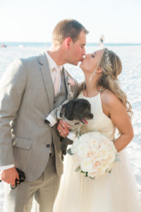 Outdoor Beach Wedding Bride and Groom Portrait With Dog wearing Mini Gray Suit, Groom in Gray Suit with BLush PInk Tie and Rose Boutonniere, Bride in Halter Ballgown Hayley Paige Wedding Dress with White Rose Bouquet and Jeweled Headband | Whimsical Clearwater BeachWedding
