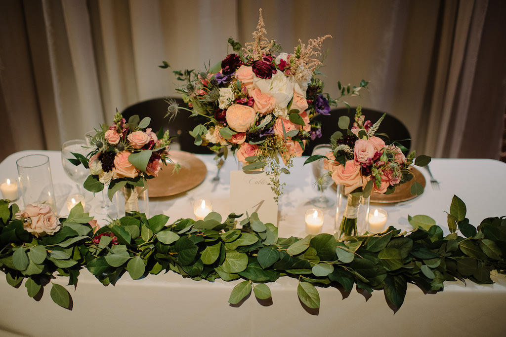 Wedding Reception Sweetheart Table with Greenery Garland, and Peach Rose, Pink, White, Magenta, and Purple Flowers with Greenery Low Centerpiece in Glass Vase with Gold Chargers