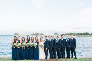 Outdoor Wedding Party Portrait in Navy Blue Cross Halter Lulus.com Dresses, Enzoani Sweetheart Lace wedding Dress, with White and Greenery Bouquets, Groomsmen in Blue Suits with Brown Shoes and White Boutonniere | Waterfront Downtown St Pete Wedding Portrait