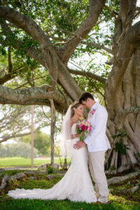 Outdoor Garden Bride and Groom Portrait, Bride in Strapless Lace Essense of Australia Wedding Dress, Groom in White Suit Jacket with Tan Pants Pink Boutonniere, With Red and White Tropical Bouquet | Tampa Bay Beach Wedding Venue Longboat Key Club