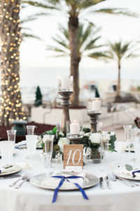 Outdoor Waterfront Wedding Reception Round Table with White Painted on Wood Table Number, Wooden Pillar Candle Holders, and Low White Rose and Greenery Centerpieces, Napkins in Blue Ribbon | Tampa Bay Wedding Venue The Westshore Yacht Club | Planner Unique Weddings and Events