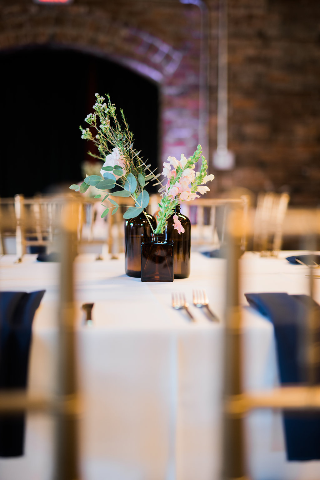 Industrial Chic Wedding Reception with Navy Blue Napkins, Gold Chiavari Chairs, Round Tables and Vintage Brown Glass Bottle Centerpiece with Natural Greenery and Pink Florals | Tampa Bay Historic Architecture Wedding Venue CL Space | Linen Rentals and Catering Tastes of Tampa Bay