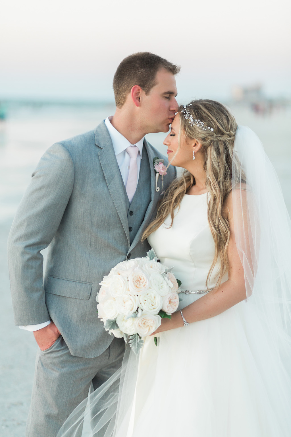 Outdoor Beach Wedding Bride and Groom Portrait, Groom in Gray Suit with Blush Pink Tie and Rose Boutonniere, Bride in Halter Ballgown Hayley Paige Wedding Dress with White Rose Bouquet and Jeweled Headband | Whimsical Clearwater Beach Wedding