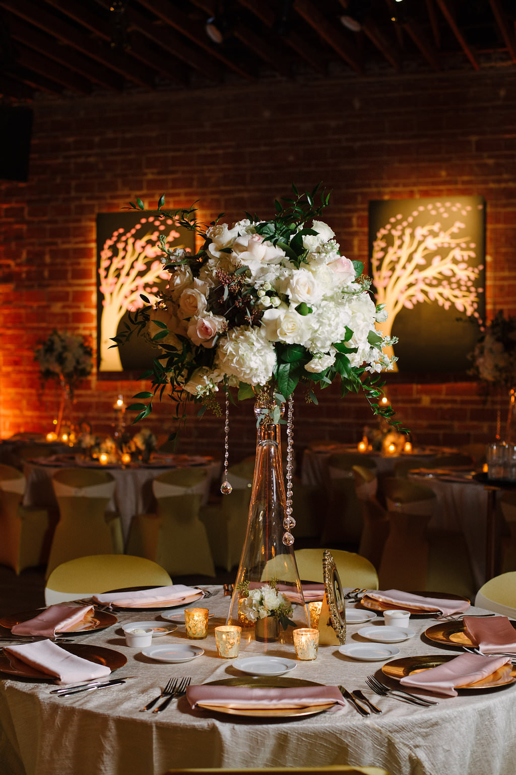 Gold and Pink Wedding Reception with Round Tables with Satin Linens, Gold Fabric Covered Chairs, Extra Tall Pink FLoral and Greenery Centerpieces in Glass Vases with Hanging Crystals