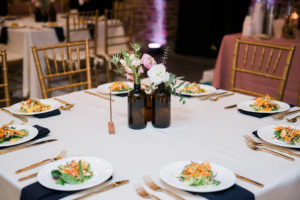 Industrial Chic Wedding Reception with Navy Blue Napkins, Gold Chiavari Chairs, Square Tables and Vintage Brown Glass Bottle Centerpiece with Natural Greenery and Pink Florals | Tampa Bay Historic Architecture Wedding Venue CL Space | Linen Rentals and Catering Tastes of Tampa Bay