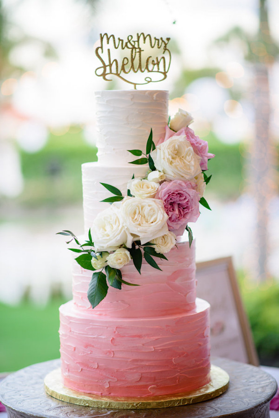 Four Tier Round White to Pink Ombre Wedding Cake on Gold Flat Cake Stand with Stylish Gold Caketopper and White and Pink Roses with Greenery