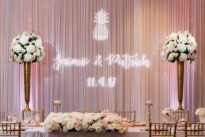 Pink and Gold Hotel Ballroom Wedding Reception With Custom Pineapple Projection on Pink Draping, Tall White Hydrangea, Pink Rose, and Greenery Centerpiece in Tall Gold Vases, Low Centerpiece with Stylish Gold Candlesticks, and Gold Chiavari Chairs | Tampa Bay Wedding Draping and Furniture Rentals Gabro Event Services
