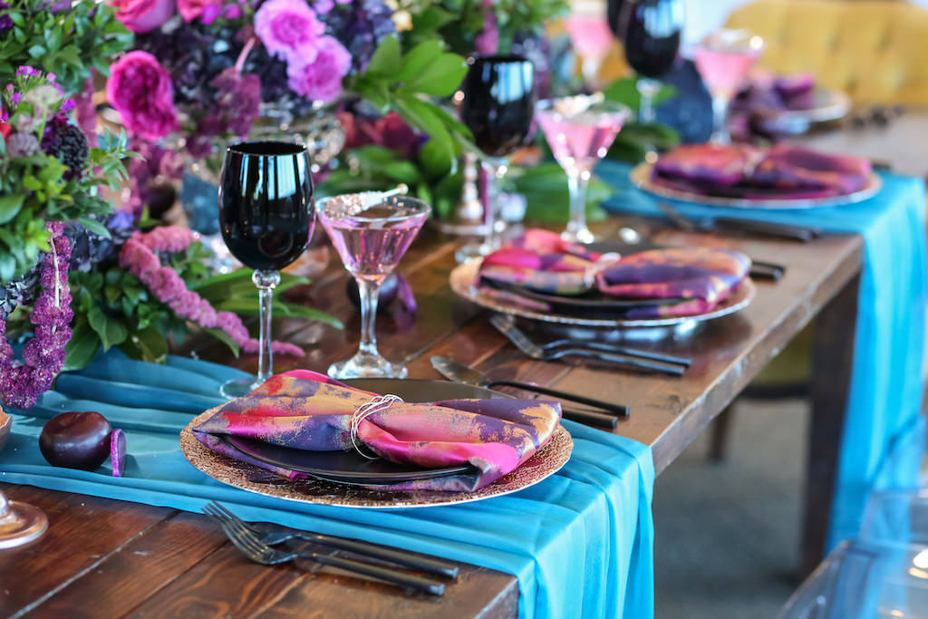 Vibrant Whimsical Wedding Reception Wooden Feasting Table with Black Flatware, Wine Glasses, and Plates, Copper Chargers and Votive Candleholders, Low Plum and Fuchsia Florals with Greenery Centerpieces, Pink and Purple Watercolor Napkins and Turquoise Linen Runners, Glitter Rock Candy Favors | Tampa Bay Wedding Planner Kelly Kennedy Weddings and Events | Furniture Rentals A Chair Affair | Florist Gabro Event Services