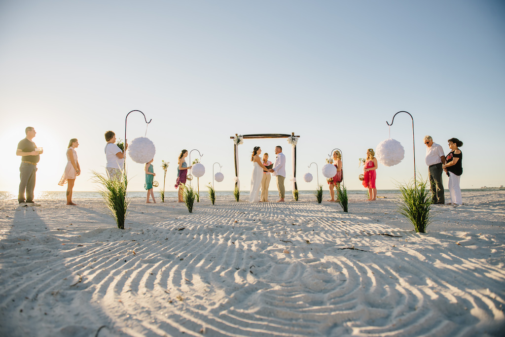 Simple Intimate Beach Wedding Ceremony Portrait with White Pomander Balls on Iron Hook with Greenery, Black Tulle Draped Table, Wooden Arch with White Flowers and Draping, and Tiki Torches, Bridesmaids in Mismatched Bright Colored Tropical Dresses | Tampa Bay Wedding Planner Gulf Beach Weddings | Treasure Island Venue Sunset Vista Condo Hotel Resort 