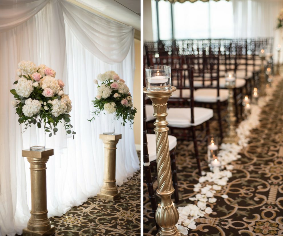Indoor Hotel Wedding Ceremony Decor with White Hydrangea and Blush Pink Rose with Greenery Flowers in Glass Vases on Ornate Gold Pillars, and Floating Votive Candles on Gold Pedestals with Petal Aisle and Wooden Chiavari Chairs with White Cushions