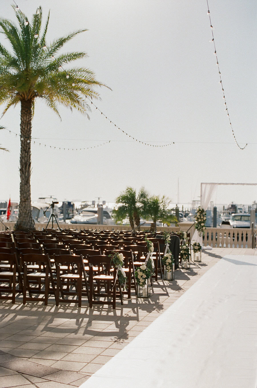 Outdoor Waterfront Wedding Ceremony with Wooden Folding Chairs, White and Greenery Florals, and White Draped Ceremony arch and String Lights | Tampa Bay Wedding Venue Westshore Yacht Club | Planner Unique Weddings and Events