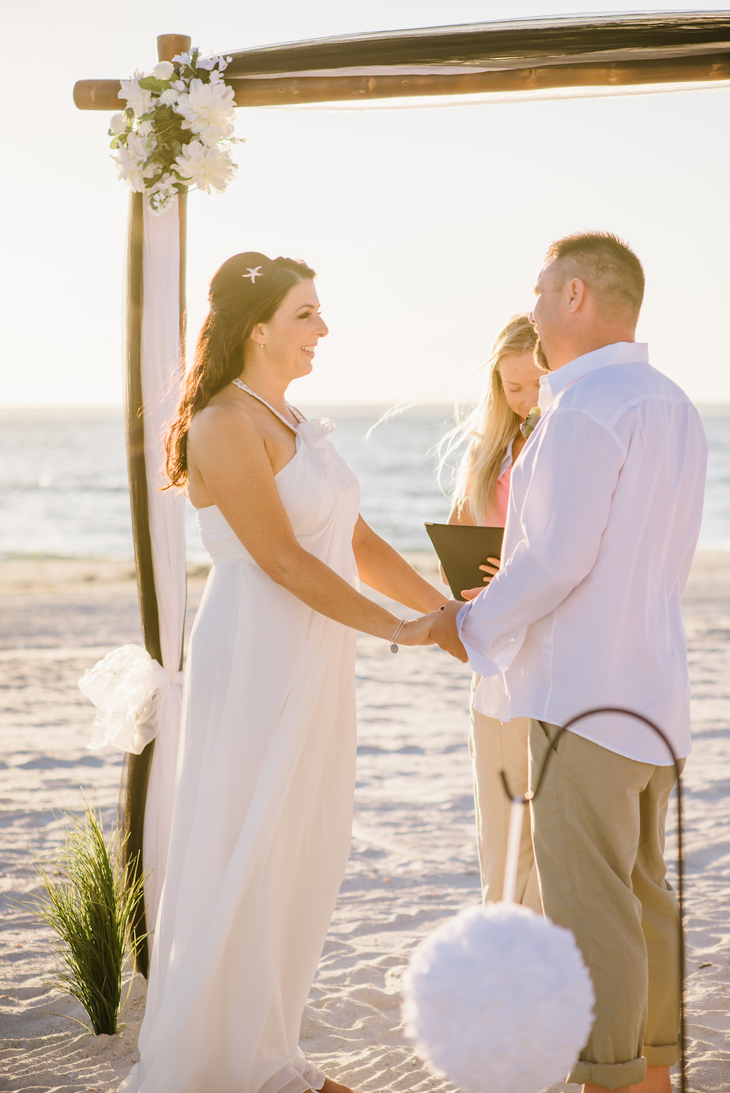 Outdoor Beach Wedding Ceremony with White Draped Wooden Ceremony Arch with Flowers and Greenery, Bride in Halter DaVinci Dress | Tampa Bay Officiant and Planner Gulf Beach Weddings