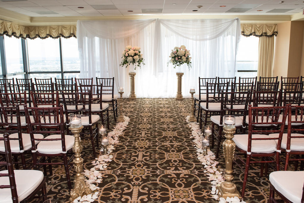 Indoor Hotel Wedding Ceremony Decor with White Hydrangea and Blush Pink Rose with Greenery Flowers in Glass Vases on Ornate Gold Pillars, and Floating Votive Candles on Gold Pedestals with Petal Aisle and Wooden Chiavari Chairs with White Cushions | Downtown Tampa Wedding Venue The Tampa Club