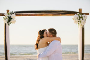 Outdoor Treasure Island Beach Wedding Ceremony First Kiss Portrait with White Draped Wooden Ceremony Arch with Flowers and Greenery