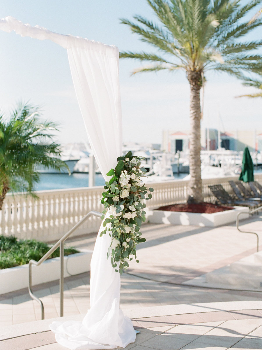 Outdoor Waterfront Wedding Ceremony with White Draped Arch with White Roses and Natural Greenery | Tampa Bay Wedding Planner Unique Weddings and Events | Venue The Westshore Yacht Club