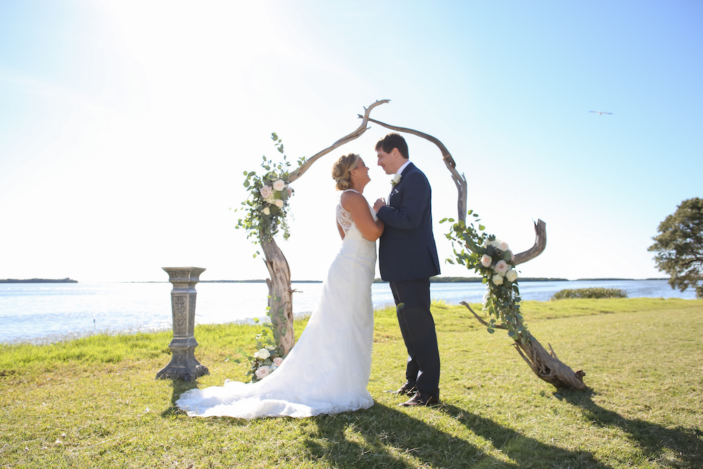 Outdoor Waterfront Wedding Ceremony Portrait with Organic Driftwood Arch with White Rose and Natural Greenery Florals, Bride in Lace Open Back Column Dress with Train | Venue Tampa Bay Watch | St Petersburg Wedding Photographer Lifelong Photography Studio