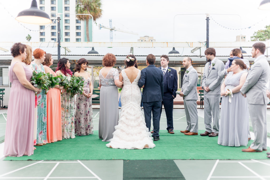 Outdoor Wedding Ceremony Portrait, Bride in Open Backed A Line Fringe David's Bridal Dress, Groomsmen in Gray Suits, Groom in Navy Blue, Bridesmaids in Mismatched Pastel Floral Print Dresses with Greenery Bouquets | Unique Historic Downtown St Pete Wedding Venue St Petersburg Shuffleboard Club