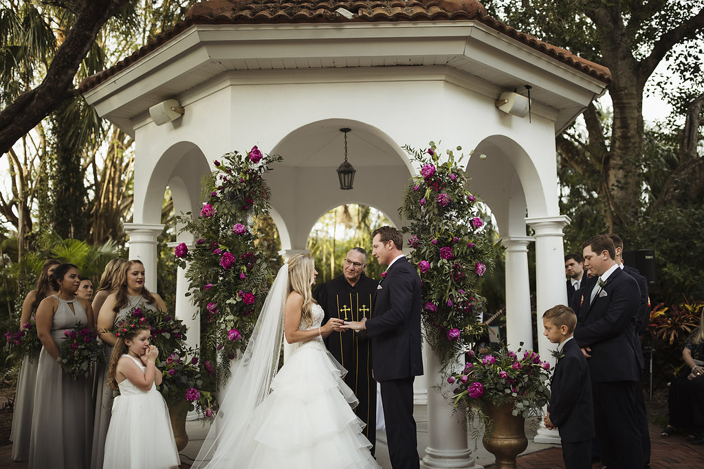 Outdoor Tropical Garden Gazebo Wedding Ceremony Portrait with Fuchsia and Pink Floral with tropical Greenery, Large Gold Classical Planters, Bride in Layered Ballgown Dress, Flower Girl with Floral Crown, Bridesmaids in Mismatched Gray Azazie Dresses | Venue Longboat Island Chapel | Sarasota Wedding Planner Jennifer Matteo Event Planning