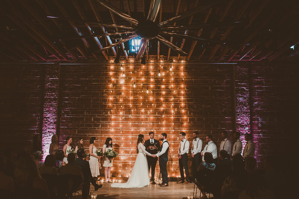 Dramatic Minimalist Indoor Wedding Ceremony Portrait with Hanging Edison Bulb String Light Backdrop on Exposed Brick, Bridesmaids in Blush Pink with Greenery Bouquets, Groomsmen in White Shirts with Suspenders and Bow Ties, Groom in Navy Blue Vest | Unique Downtown St Pete Wedding Venue NOVA 535