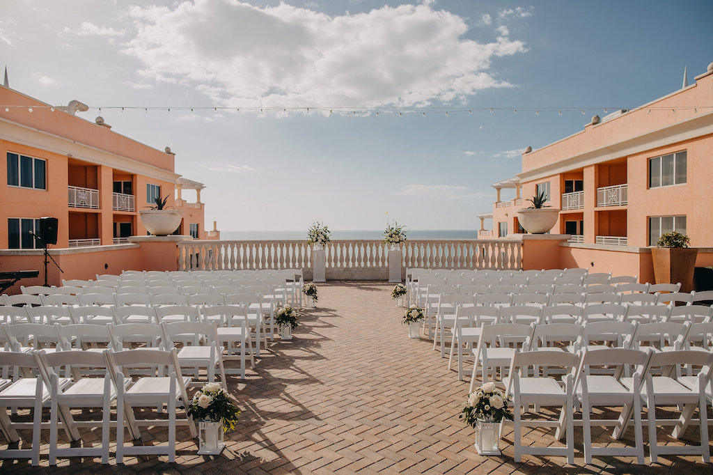Waterfront Tampa Bay Luxury Hotel Wedding Ceremony with White Folding Chairs, Ivory Florals with Greenery in Hurricane Lanterns in Aisle and on Pedestals | Tampa Bay Wedding Venue Hyatt Clearwater Beach and Spa