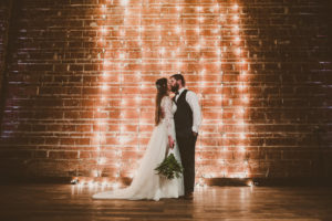 Dramatic Minimalist Indoor Wedding Ceremony First Kiss Portrait with Hanging Edison Bulb String Light Backdrop on Exposed Brick, Bride in Long Sleeve Davids Bridal Dress with Green Fern Bouquet, Groom in Navy Blue Vest and Bow Tie | Unique Downtown St Pete Wedding Venue NOVA 535