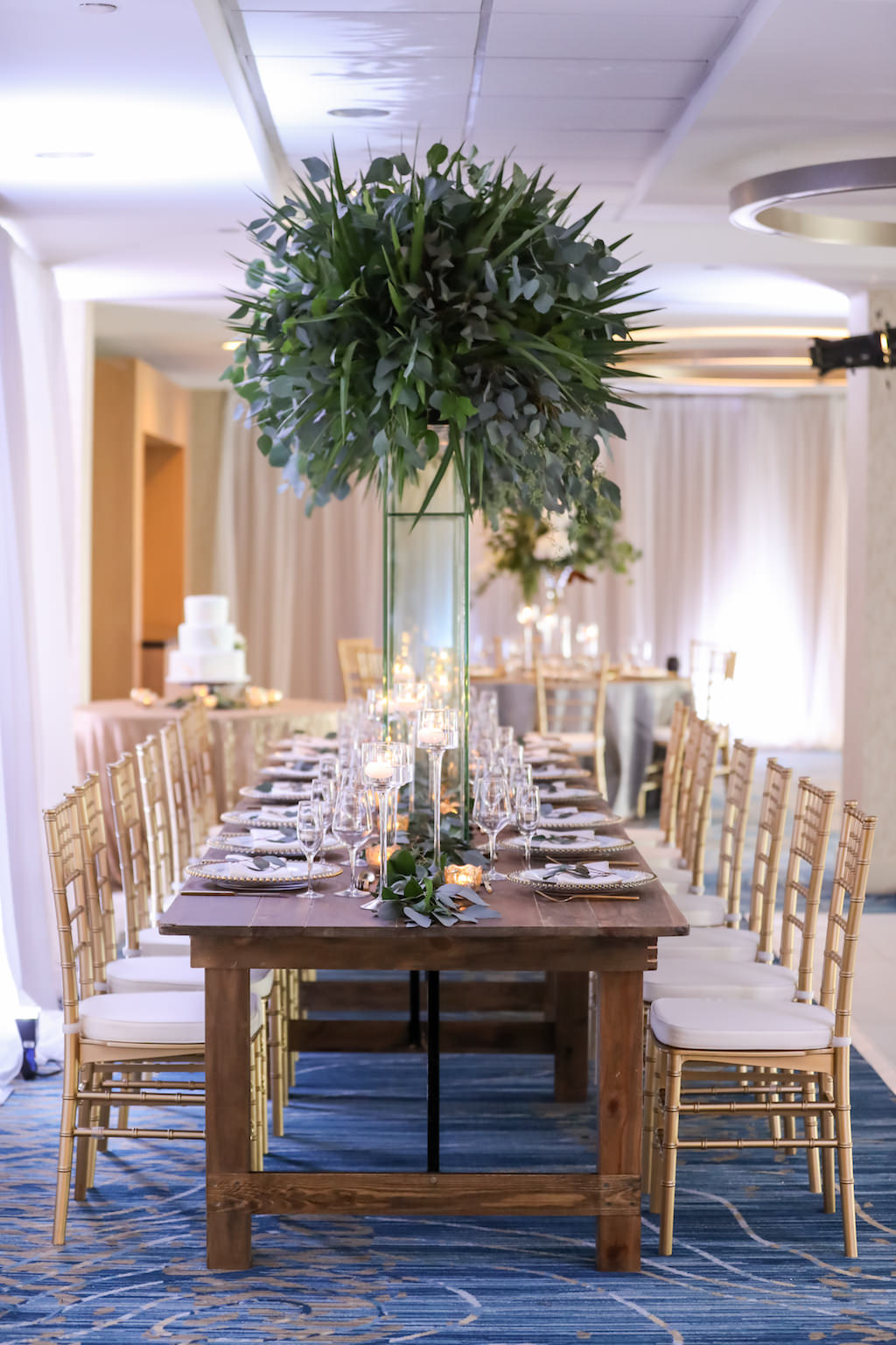 Hotel Ballroom Wedding Reception With Long Wood Feasting Table, Extra Tall Greenery Centerpiece in Clear Glass Rectangular Vases, Gold Chiavari Chairs, White Draping, and Glass Candleholders at Varying Heights and Gold Beaded Glass Chargers | Beach Hotel Wedding Venue Hilton Clearwater Beach