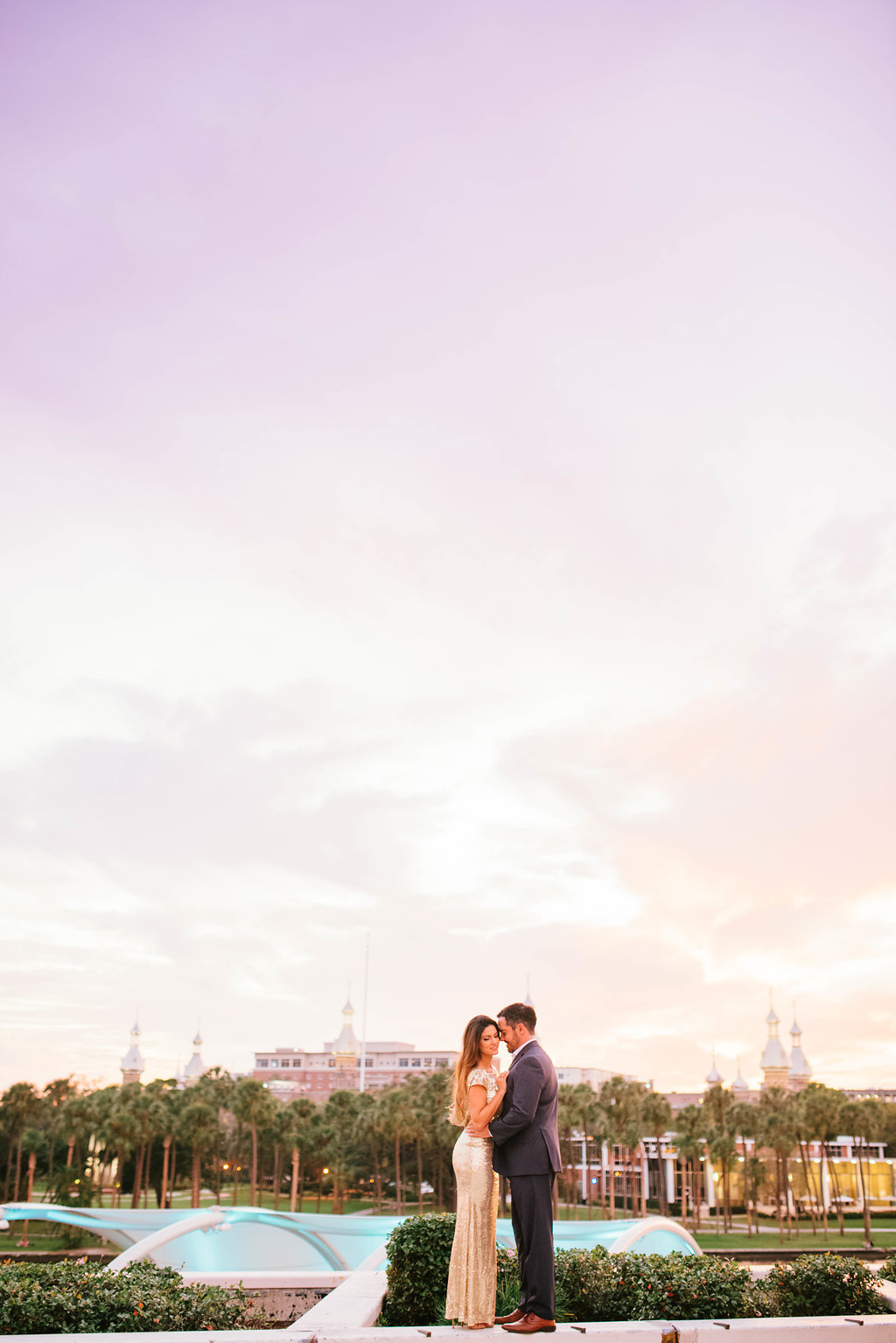 Downtown Tampa Riverwalk Classy Sunset Engagement Portrait, Bride in Long Gold Sequin Dress, Groom in Navy Blue Suit with Brown Leather Shoes with University of Tampa Skyline