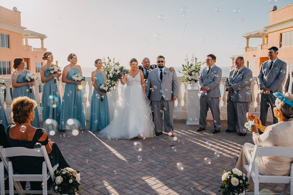 Waterfront Hotel Wedding Rooftop Ceremony Portrait, Bride in Ballgown Hayley Paige Dress, Groomsmen in Gray Suits with Black TIes, Bridesmaids in MIsmatched Light Blue Weddington Way Dresses, with White Folding Chairs, Ivory FLorals with Greenery in Hurricane Lanterns in Aisle and on Pedestals | Tampa Bay Wedding Venue Hyatt Regency Clearwater Beach and Spa | Photographer Rad Red Creative
