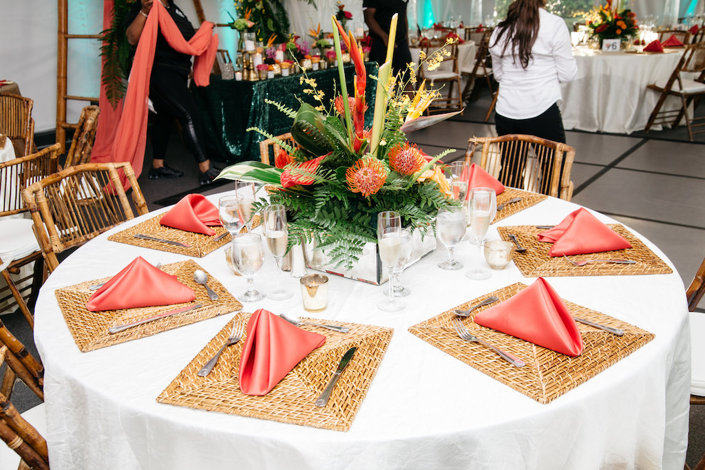 Tented Waterfront Wedding Reception with Low Bird of Paradise Orange and Yellow Floral with Tropical Greenery Centerpiece in Square Glass Vase, with Guava Pink Napkins and Rattan Folding Chairs, Square Bamboo Placemats | Tampa Bay Wedding Venue DoubleTree Suites by Hilton