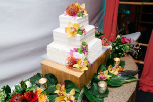 Three Tier Square White Wedding Cake on Silver and Bamboo Cake Stand with Orange, Guava, Purple and Yellow Tropical Flowers with Greenery on Sequin Linen with Votive Candles