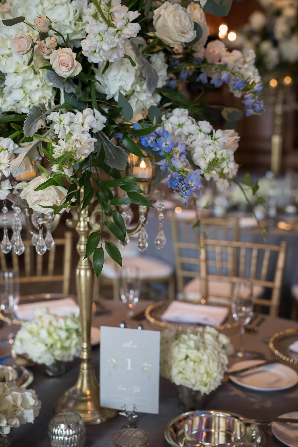 Elegant Wedding Reception Table with Tall White, Blush and Blue Floral with Greenery and Crystals Centerpiecee in Gold Candelabra, with Small Hydrangea Flowers, Gray Printed Table Number and Silk Linen, and Gold Chiavari Chairs