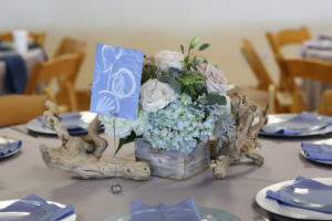 Ocean Inspired Blue Wedding Reception Low Birchwood, Blush Rose and Organic Greenery in Wooden Box Centerpiece with Aquatic Blue Printed Paper Table Number, Blue Napkins on Silver Chargers